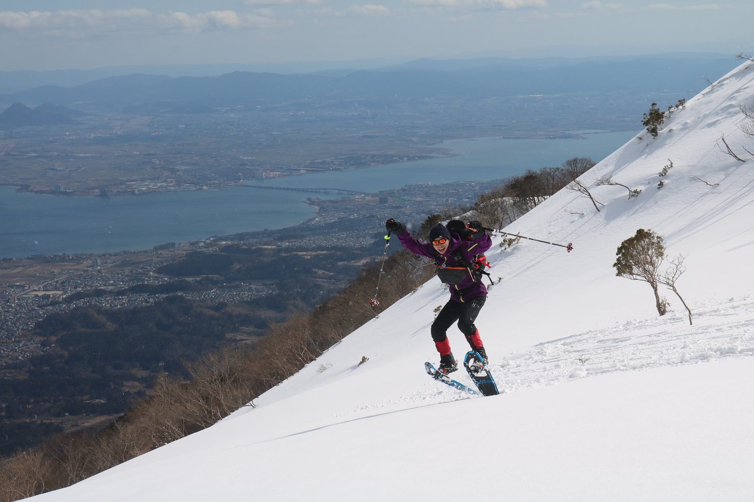 雪の急斜面を登る筆者（谷本）と、眼下に広がる湖と街並み